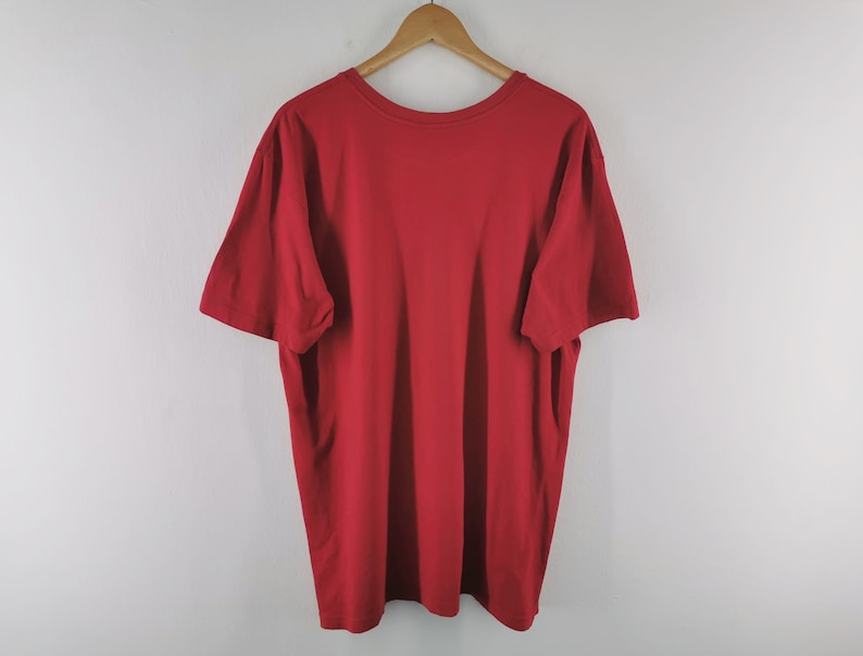 May include: A solid red t-shirt with a crew neck and short sleeves hangs from a wooden hanger. The shirt is made of a soft material and is displayed against a plain white background. The t-shirt is a basic, casual garment.