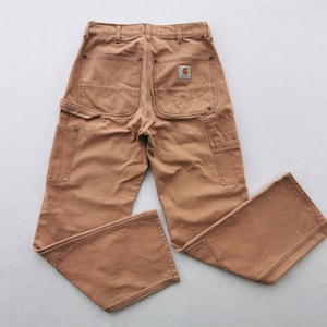 May include: Tan work pants with multiple pockets and a Carhartt logo patch on the back. The pants have a classic workwear design with reinforced stitching and a relaxed fit. The color is a warm, earthy tone.