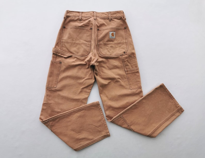 May include: Tan work pants with multiple pockets and a Carhartt logo patch on the back. The pants have a classic workwear design with reinforced stitching and a relaxed fit. The color is a warm, earthy tone.