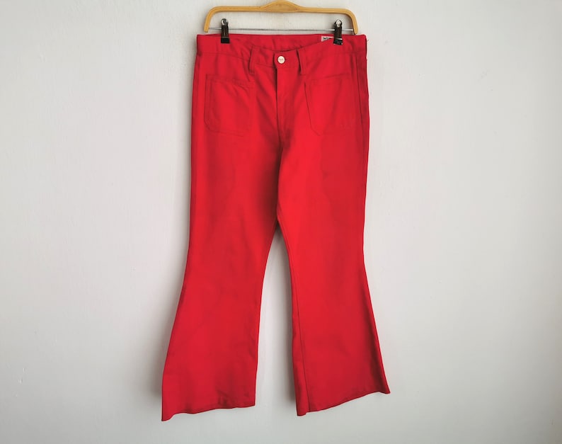 May include: Bright red flared pants hanging on a wooden hanger. The pants have two front pockets and a button closure. The fabric appears to be a solid color, with a slight sheen. The pants are a bold, retro style.