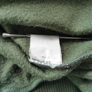 May include: Close-up of green fabric with a white tag held by a silver pin. The tag has Japanese text. The fabric has a textured, fuzzy appearance.