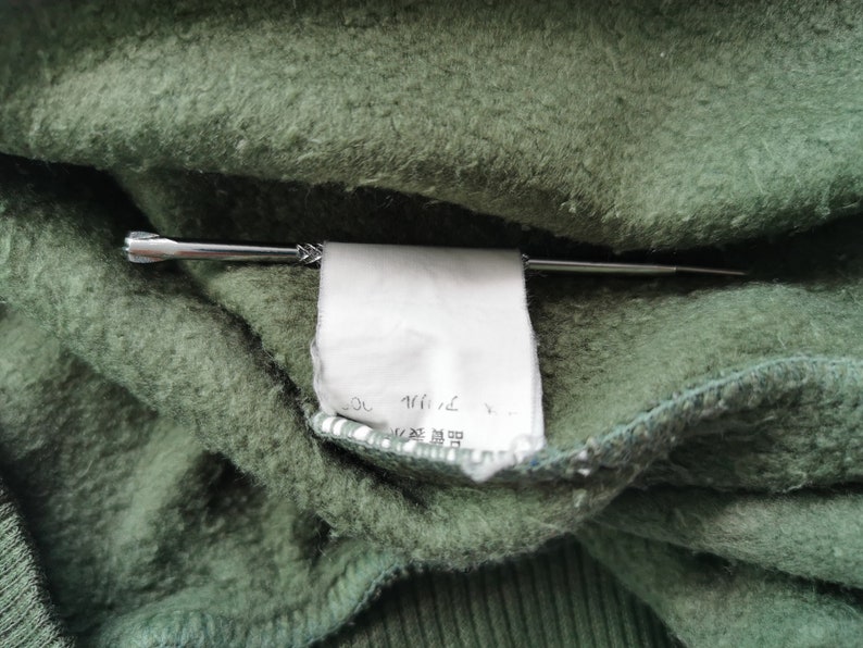 May include: Close-up of green fabric with a white tag held by a silver pin. The tag has Japanese text. The fabric has a textured, fuzzy appearance.