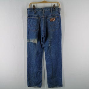 May include: Vintage blue denim jeans with a classic five-pocket design. The jeans have a faded appearance and a patch on the left leg. A brown leather patch is on the back pocket. The jeans are hanging on a wooden hanger.