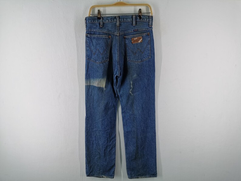 May include: Vintage blue denim jeans with a classic five-pocket design. The jeans have a faded appearance and a patch on the left leg. A brown leather patch is on the back pocket. The jeans are hanging on a wooden hanger.