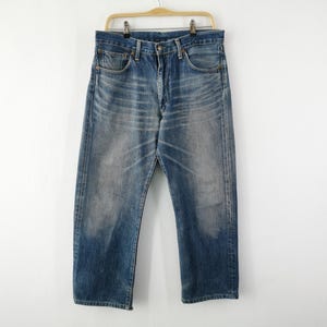 May include: Faded blue denim jeans hanging on a wooden hanger. The jeans have a classic five-pocket design and a straight leg cut. The denim has a worn, distressed look with lighter areas.