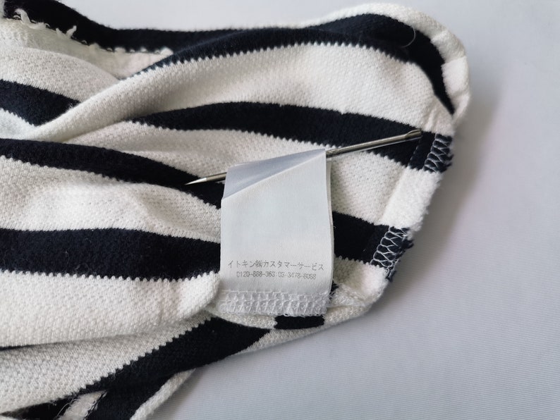 Puede incluir: Primer plano de una tela a rayas blancas y azul marino. La tela est&aacute; doblada, con una etiqueta sujeta con un alfiler. La etiqueta tiene texto japon&eacute;s. La tela parece ser de punto.