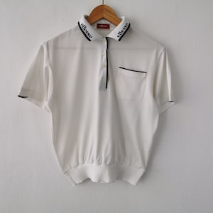 May include: White polo shirt with black trim and the brand name "ellesse" on the collar. The shirt has short sleeves, a pocket, and a button-up placket. The shirt is hanging on a wooden hanger.