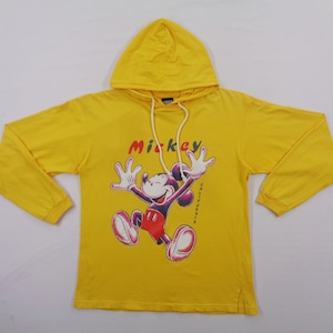 Op de afbeelding: Gele hoodie met een Mickey Mouse-afbeelding. Het woord "Mickey" staat boven het personage en "California" is verticaal aan de rechterkant afgedrukt. De sweatshirt heeft lange mouwen en een capuchon met trekkoord.
