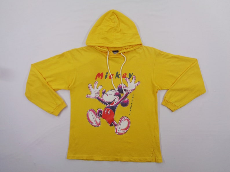 Op de afbeelding: Gele hoodie met een Mickey Mouse-afbeelding. Het woord "Mickey" staat boven het personage en "California" is verticaal aan de rechterkant afgedrukt. De sweatshirt heeft lange mouwen en een capuchon met trekkoord.