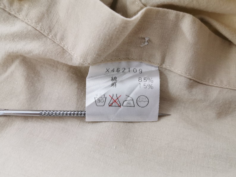 May include: A beige fabric garment tag featuring the text X462109, 85% cotton, and 15% linen, indicated by Japanese characters. The white rectangular label includes four black laundry care symbols, with one symbol marked by a red cross.