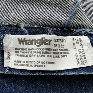 May include: Close-up of a Wrangler jeans label. The label is white with black text, including the brand name, size 36 x 32, and care instructions. The jeans are dark blue denim.