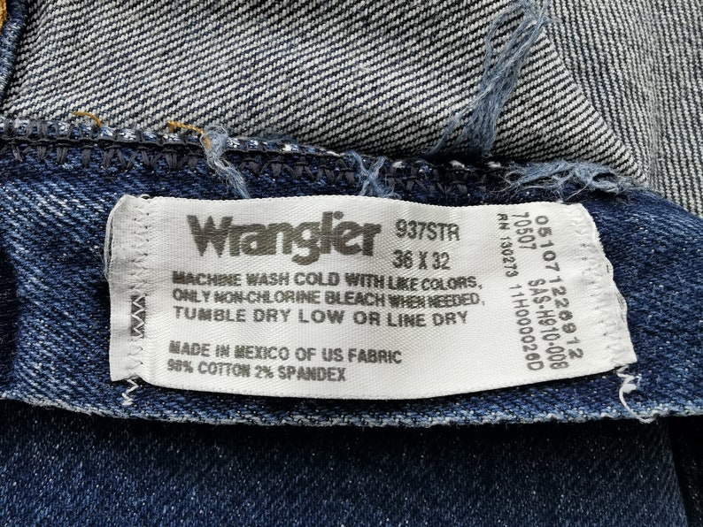 May include: Close-up of a Wrangler jeans label. The label is white with black text, including the brand name, size 36 x 32, and care instructions. The jeans are dark blue denim.