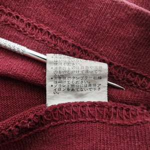 May include: Close-up of a burgundy fabric with a stitched hem and a small tag. A silver tool is placed on the fabric. The tag has Japanese text with care instructions.