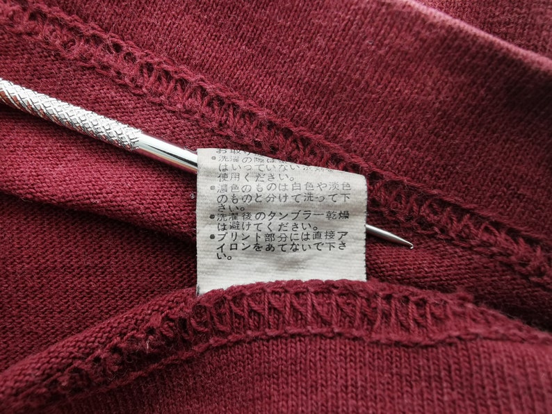 May include: Close-up of a burgundy fabric with a stitched hem and a small tag. A silver tool is placed on the fabric. The tag has Japanese text with care instructions.