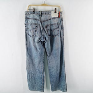 May include: Light blue denim jeans hanging on a wooden hanger. The jeans have a classic five-pocket design, with a faded wash and visible wear marks. The back pockets feature a distinctive stitching pattern.
