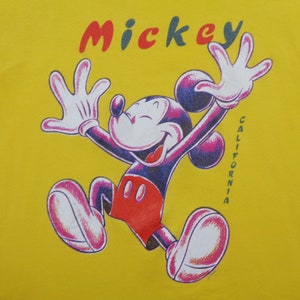 Op de afbeelding: Geel t-shirt met een afbeelding van Mickey Mouse in een vrolijke pose, armen omhoog. Het woord "Mickey" staat in rood, blauw en groen boven het personage. Het woord "CALIFORNIA" staat verticaal aan de rechterkant.