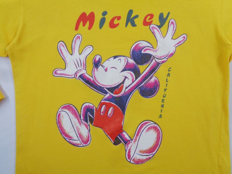 Op de afbeelding: Geel t-shirt met een afbeelding van Mickey Mouse in een vrolijke pose, armen omhoog. Het woord "Mickey" staat in rood, blauw en groen boven het personage. Het woord "CALIFORNIA" staat verticaal aan de rechterkant.