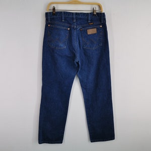 K&ouml;nnte beinhalten: Dunkelblaue Jeans aus Denim, die an einem Holzb&uuml;gel h&auml;ngen. Die Jeans haben ein klassisches F&uuml;nf-Taschen-Design, Kupfernieten und einen braunen Lederaufn&auml;her auf der R&uuml;ckseite. Das Wrangler-Logo ist sichtbar.