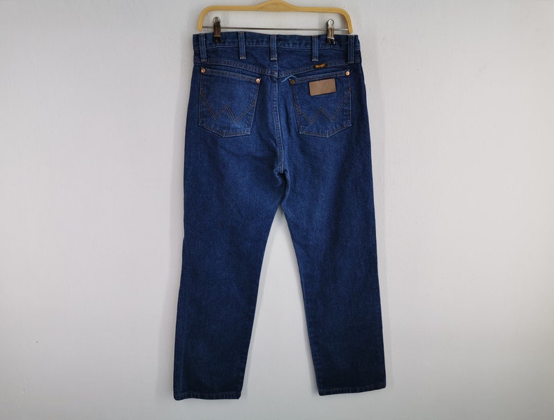 K&ouml;nnte beinhalten: Dunkelblaue Jeans aus Denim, die an einem Holzb&uuml;gel h&auml;ngen. Die Jeans haben ein klassisches F&uuml;nf-Taschen-Design, Kupfernieten und einen braunen Lederaufn&auml;her auf der R&uuml;ckseite. Das Wrangler-Logo ist sichtbar.