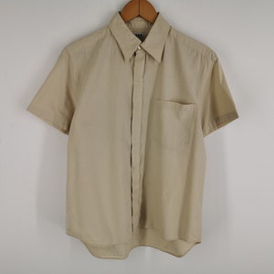 May include: A short-sleeved button-down shirt in a solid beige color. The shirt features a classic pointed collar, a single chest pocket on the left side, and a buttoned front placket. The garment is displayed on a wooden hanger against a plain background.