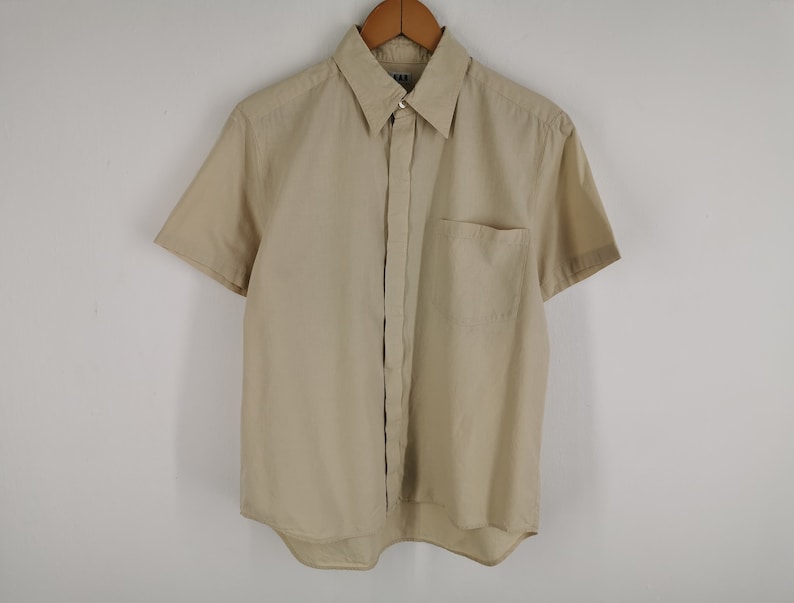 May include: A short-sleeved button-down shirt in a solid beige color. The shirt features a classic pointed collar, a single chest pocket on the left side, and a buttoned front placket. The garment is displayed on a wooden hanger against a plain background.