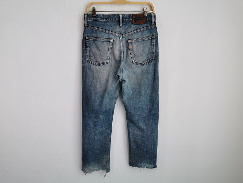 Puede incluir: Vaqueros azules descoloridos con un aspecto desgastado, colgados de una percha de madera. Los vaqueros tienen un dise&ntilde;o cl&aacute;sico de cinco bolsillos, un parche de cuero y una etiqueta roja. Los dobladillos inferiores est&aacute;n deshilachados.