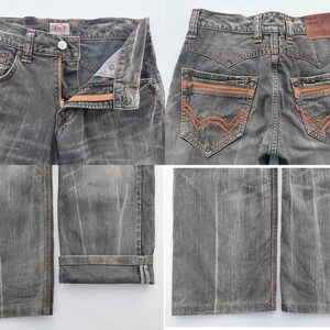 May include: A pair of gray denim jeans with orange stitching and zipper details. The jeans have a classic five-pocket design, a button and zipper closure, and a leather patch on the back. The jeans are folded at the bottom.