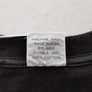 May include: Close-up of a black t-shirt with a white tag attached. The tag has text that reads "MACHINE WASH COLD WATER DO NOT TUMBLE DRY 100% COTTON". The shirt is made of cotton.
