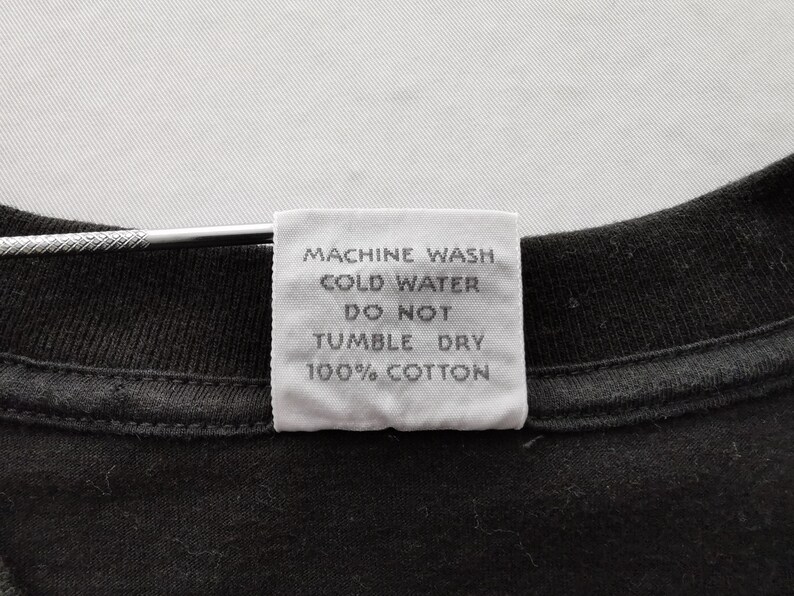 May include: Close-up of a black t-shirt with a white tag attached. The tag has text that reads "MACHINE WASH COLD WATER DO NOT TUMBLE DRY 100% COTTON". The shirt is made of cotton.