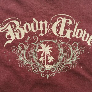 May include: Maroon t-shirt with the text "Body Glove" in a decorative, vintage style. Below the text is a circular design with palm trees and decorative flourishes in a pale green colour.
