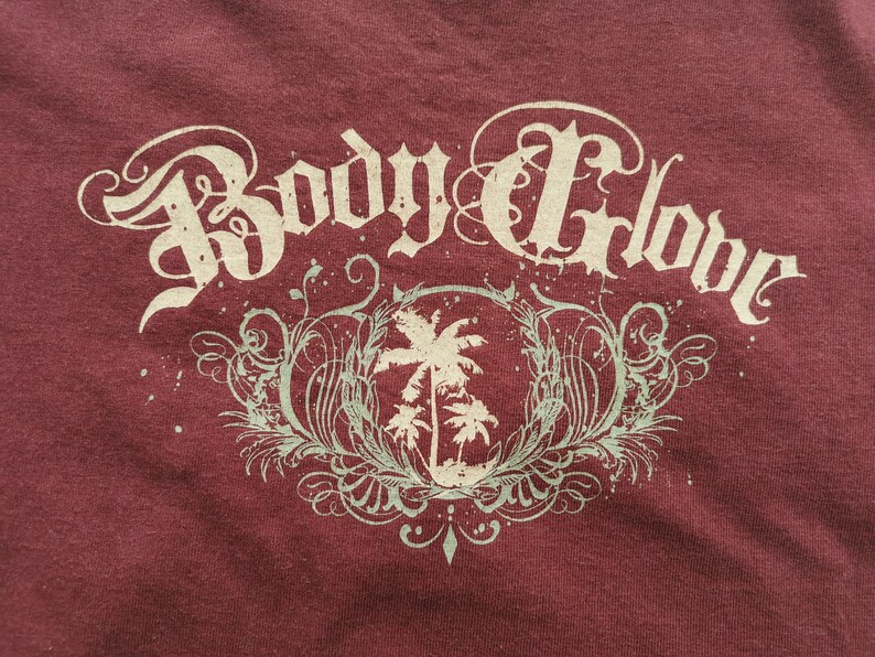 May include: Maroon t-shirt with the text "Body Glove" in a decorative, vintage style. Below the text is a circular design with palm trees and decorative flourishes in a pale green colour.