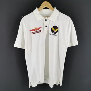 May include: White polo shirt with short sleeves, featuring patches with the text "ALPHA INDUSTRIES" and "U.S. AIR FORCE". The shirt has a collared neckline and button closure. The patches are red, black, and yellow.