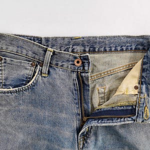 Puede incluir: Primer plano de unos vaqueros de mezclilla azul claro con la cremallera parcialmente abierta. Los vaqueros tienen un dise&ntilde;o cl&aacute;sico de cinco bolsillos, con costuras y remaches met&aacute;licos visibles. La tela muestra un aspecto descolorido y desgastado.
