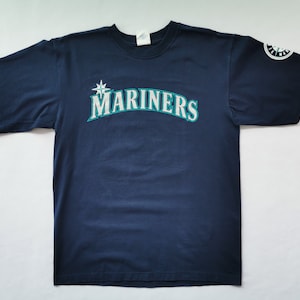 May include: Navy blue t-shirt featuring the word "MARINERS" in white and teal lettering, with a star logo. The Seattle Mariners logo is on the left sleeve. A classic baseball fan apparel item.