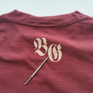 May include: A maroon t-shirt featuring the letters "BG" in a decorative, light tan font. A silver tool is placed diagonally across the shirt, pointing towards the letters. The shirt's neckline is visible at the top.
