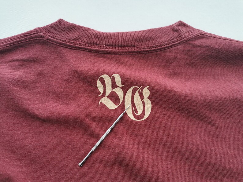 May include: A maroon t-shirt featuring the letters "BG" in a decorative, light tan font. A silver tool is placed diagonally across the shirt, pointing towards the letters. The shirt's neckline is visible at the top.