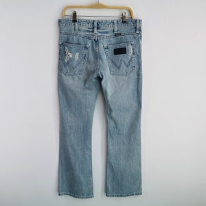 Pu&ograve; includere: Jeans di denim azzurro con un design strappato, caratterizzati da un classico stile a cinque tasche e una toppa in pelle con il logo del marchio sul retro. I jeans sono appesi a una gruccia di legno.