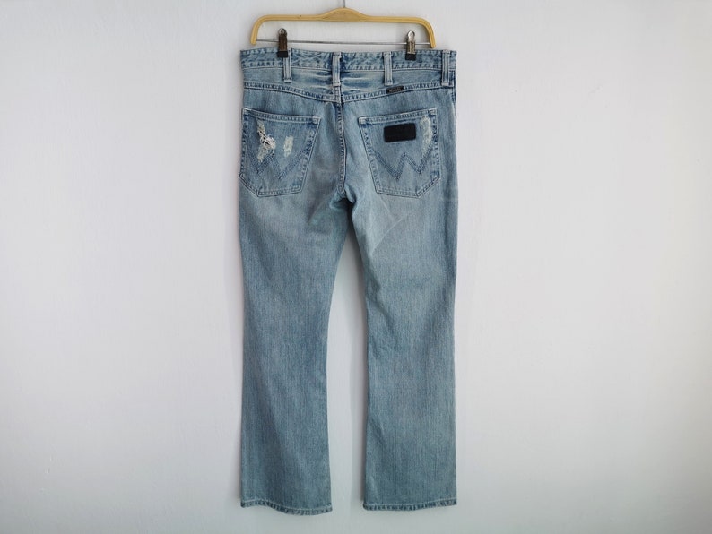 Pu&ograve; includere: Jeans di denim azzurro con un design strappato, caratterizzati da un classico stile a cinque tasche e una toppa in pelle con il logo del marchio sul retro. I jeans sono appesi a una gruccia di legno.