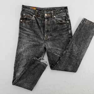 May include: High-waisted jeans in a dark acid-wash black denim featuring a button fly closure, five-pocket styling, and frayed raw-edge hems. The waistband includes a small yellow brand label with red text, and the seams are finished with copper-toned rivets.