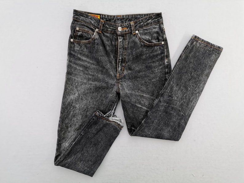 May include: High-waisted jeans in a dark acid-wash black denim featuring a button fly closure, five-pocket styling, and frayed raw-edge hems. The waistband includes a small yellow brand label with red text, and the seams are finished with copper-toned rivets.