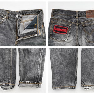 May include: Acid-washed black denim jeans featuring a button fly, a brown leather EDWIN brand patch on the back waistband, and a rectangular patch on the back pocket with the red text SCORPIONS. The jeans have frayed hems and orange stitching.