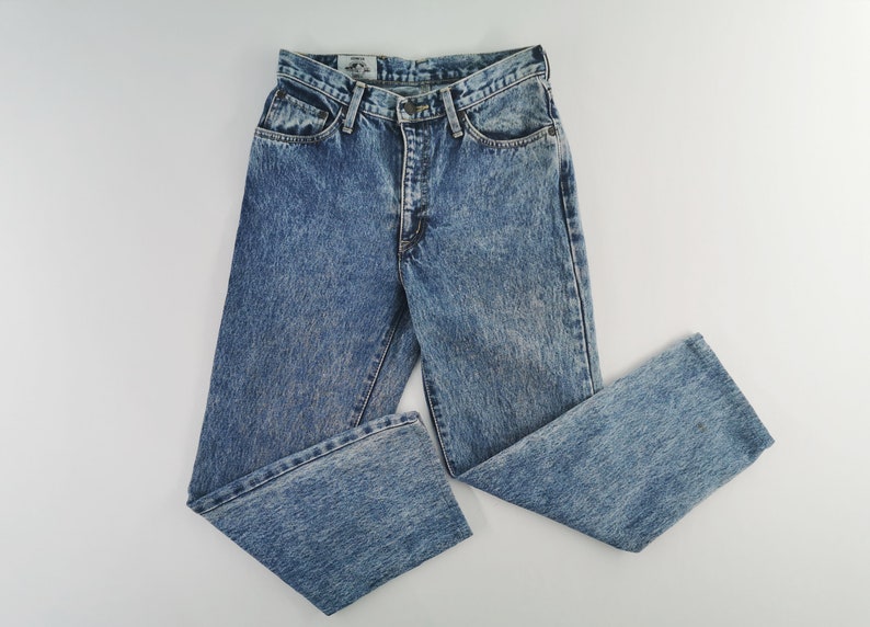 Puede incluir: Vaqueros azul claro con lavado &aacute;cido y dise&ntilde;o cl&aacute;sico de cinco bolsillos. Los pantalones vaqueros tienen cierre de bot&oacute;n y cremallera y un corte recto. Los vaqueros est&aacute;n colocados sobre una superficie blanca.