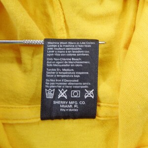 Op de afbeelding: Close-up van een geel kledingstuk met een zwart etiket. Het etiket bevat wasvoorschriften in meerdere talen, waaronder Engels, Frans en Spaans. Het etiket bevat ook de fabrikantgegevens: SHERRY MFG. CO., MIAMI, FL, RN #16490.