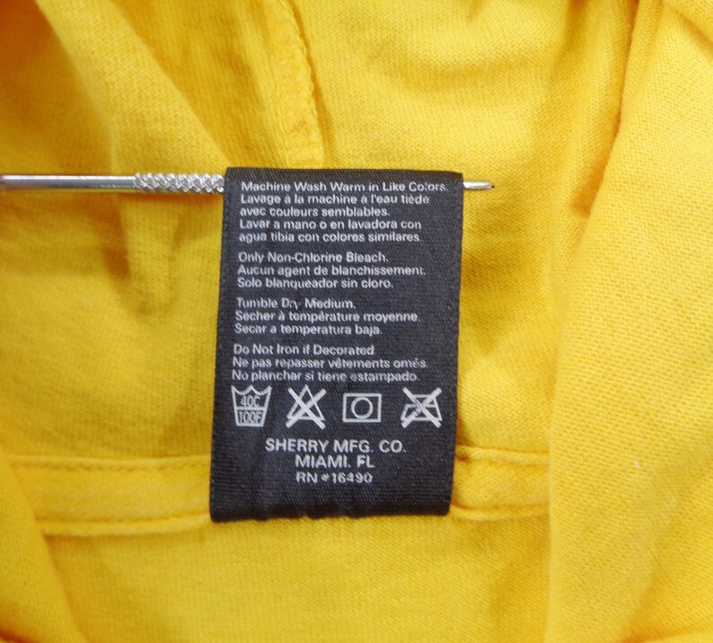 Op de afbeelding: Close-up van een geel kledingstuk met een zwart etiket. Het etiket bevat wasvoorschriften in meerdere talen, waaronder Engels, Frans en Spaans. Het etiket bevat ook de fabrikantgegevens: SHERRY MFG. CO., MIAMI, FL, RN #16490.