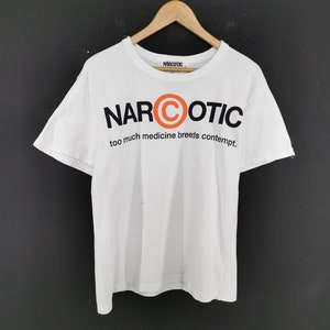 May include: White t-shirt with the word "NARCOTIC" in black and orange block letters. The shirt also features the phrase "too much medicine breeds contempt." The t-shirt is hanging on a wooden hanger.