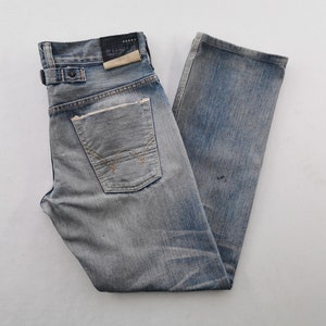 K&ouml;nnte beinhalten: Verblasste blaue Jeans aus Denim mit einem Used-Look. Merkmale sind ein Knopfverschluss, G&uuml;rtelschlaufen und eine Ges&auml;&szlig;tasche mit einzigartigem Nahtdesign. Die Jeans haben ein abgenutztes Aussehen.