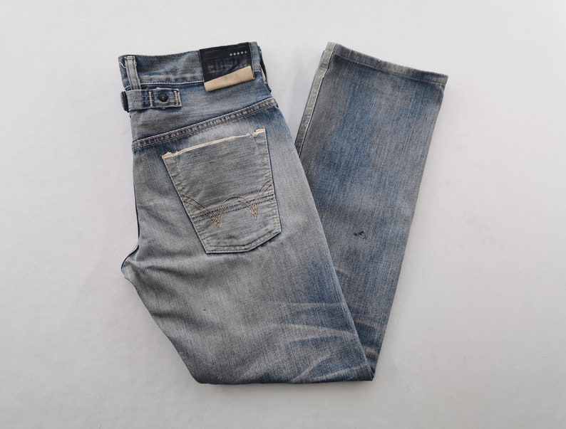 K&ouml;nnte beinhalten: Verblasste blaue Jeans aus Denim mit einem Used-Look. Merkmale sind ein Knopfverschluss, G&uuml;rtelschlaufen und eine Ges&auml;&szlig;tasche mit einzigartigem Nahtdesign. Die Jeans haben ein abgenutztes Aussehen.