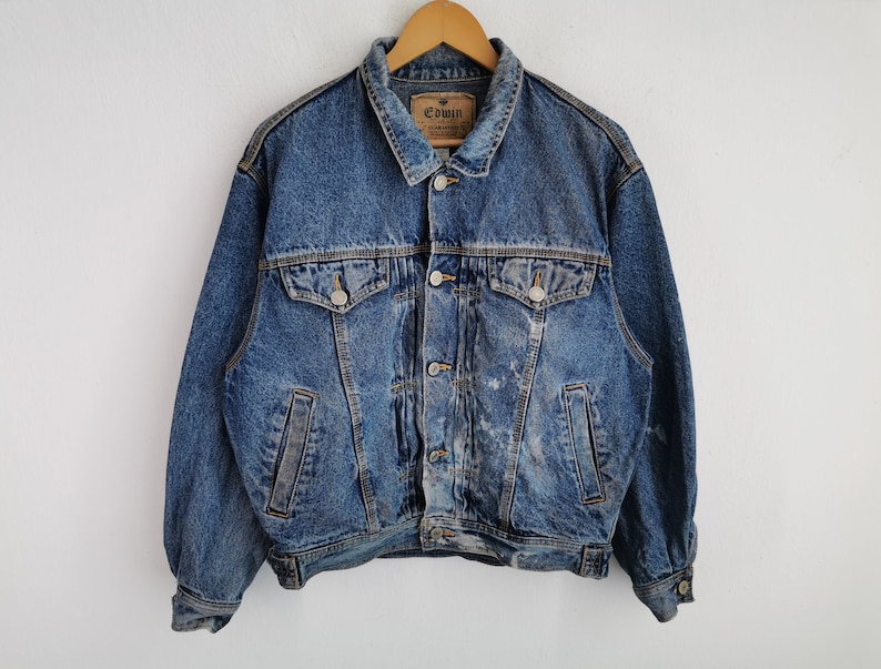 May include: A vintage blue denim jacket with a button-up front, two chest pockets, and two side pockets. The jacket has a classic collar and is hanging on a wooden hanger. The brand tag "Edwin" is visible on the collar.