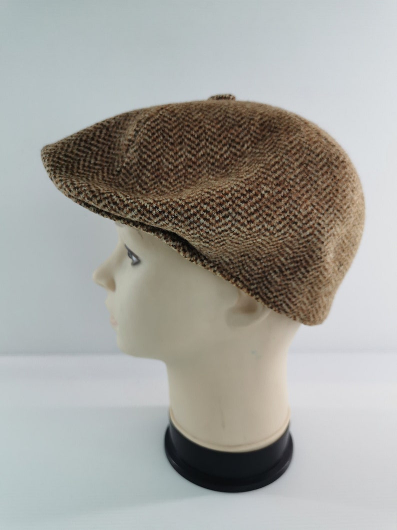 Op de afbeelding: Bruine tweed newsboy cap met een visgraatpatroon. De pet wordt getoond op een mannequinhoofd, waarbij de vorm en textuur worden benadrukt. De pet heeft een afgeronde kroon en een korte, stijve klep.