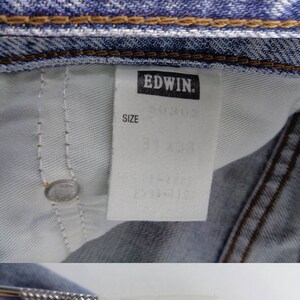 May include: Close-up of light blue denim jeans with a red and blue label that reads "JEANS 503 EDWIN." The jeans have a size tag that reads "SIZE 31 x 33." The jeans are made of 100% cotton.